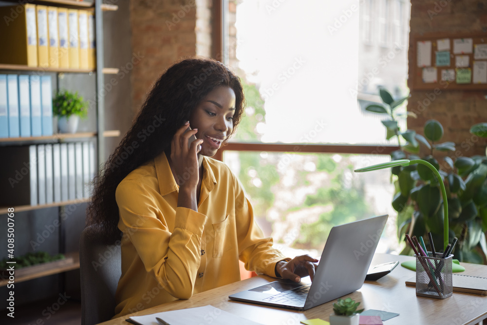 Profile side photo of young dark skin woman speak smartphone use laptop indoors in office workstation Profile side photo of young dark skin woman speak smartphone use laptop indoors in office workstation
