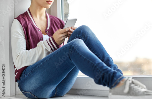 sadness and people concept - sad teenage girl with smartphone sitting on window sill