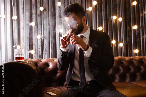 Portrait of a brutal beard man with tattoo smoking cigar in hot elegant suit  drinking whiskey alcohol in loft apartment . Couple love story concept