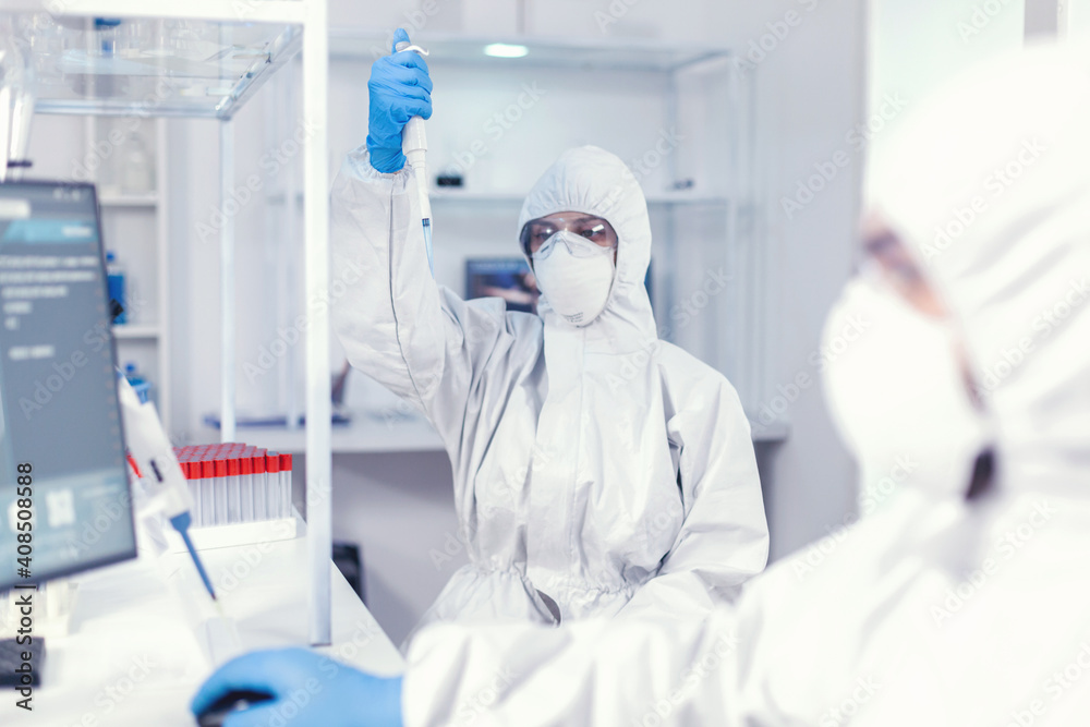 Working with micropipette using protection suit as safety precaution for coroanvirus. Team of microbiologists in research laboratory conducting experiment during global pandemic with covid19. Working with micropipette using protection suit as safety precaution for coroanvirus. Team of microbiologists in research laboratory conducting experiment during global pandemic with covid19.