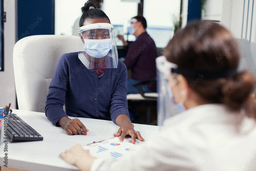 Obraz premium African woman sitting at workplace wearing face mask for covid19. Multiethnic team working in company with new normal respecting social distance because of global pandemic with coronavirus.