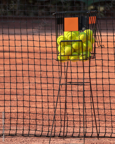 Sport tennis game concept. Tinnis balls in basket against backdrop of red clay court and net. Sports lifestyle, outdoor active leisure photo