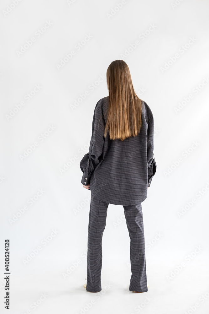Girl with long hair standing with her back in a plaid shirt and loose-fitting trousers indoors on a white background. Girl with long hair standing with her back in a plaid shirt and loose-fitting trousers indoors on a white background.
