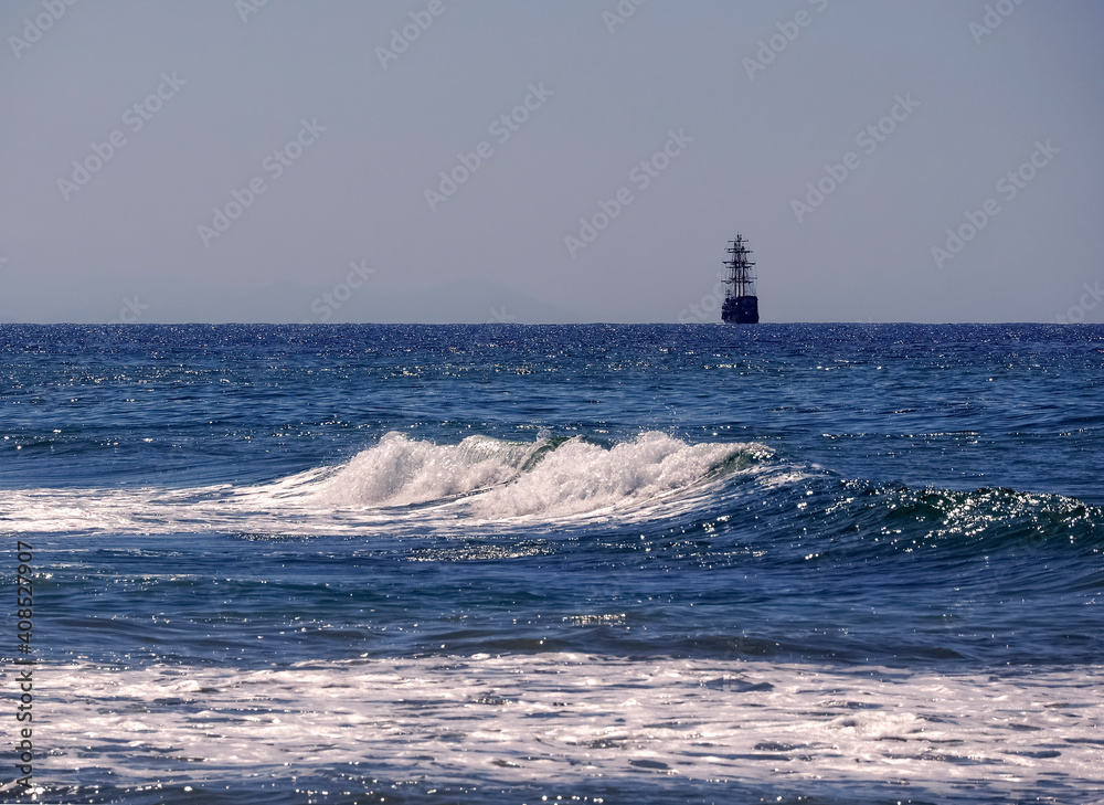 Endless blue sea. A wave rose close by. A sailing ship is visible on the horizon, the sails are lowered. Clean space for text. Natural background with sea, wave and sailboat.