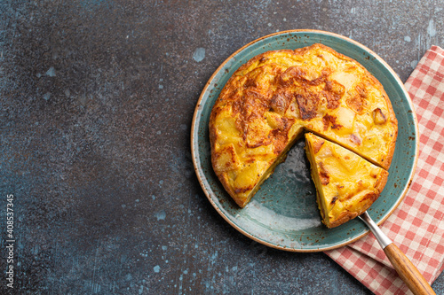 Homemade Spanish tortilla with one slice cut - omelette with potatoes on plate on stone rustic background top view. Traditional dish of Spain Tortilla de patatas for lunch or snack, space for text 