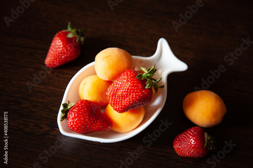 composition with a fruit plate against a dark wooden table. The fruit as a whole. Assorted apricots  strawberries iin a white deep plate in the shape of a fish. Vegetarian menu. raw food. healthy food