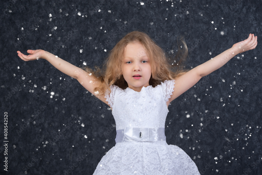 Beautiful little girl in a white dress with raised arms under flying snowflakes.