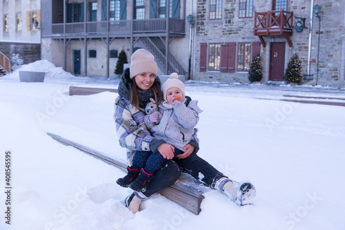 Full length horizontal shot of cute smiling young woman holding her happy toddler girl enjoying a lollipop while sitting on wooden bench in the old town during a winter afternoon, Quebec City, Quebec