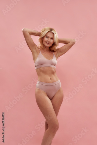 Feeling good. Happy charming middle aged caucasian blonde woman in underwear smiling at camera, keeping arms raised while standing against pink background