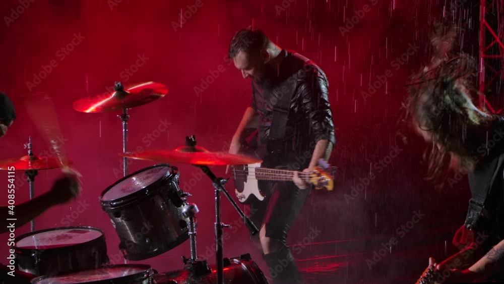 Camera slides over a group of rock musicians who play their instruments with crazy emotions in a dark studio illuminated by red lights. Drum set, electric and bass guitar. Close up. Slow motion.