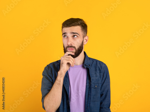 Portrait of pensive young bearded man touching his chin, deep in thought over orange studio background