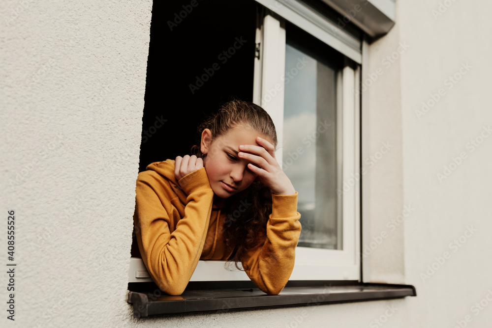 Obraz premium A little girl who is in isolation due to a coronavirus pandemic looks sadly out the window