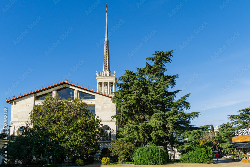 Obraz premium Sochi Commercial Sea Port with spire. Evergreen Magnolia Grandiflora and huge Himalayan Сedar (Cedrus Deodara) grow in front of famous symbol of city resort. Sochi, Russia - November 23, 2020