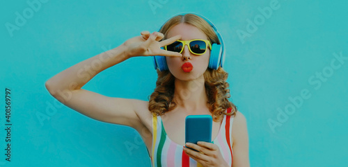Portrait of stylish young woman in wireless headphones with phone listening to music blowing her lips on a blue background
