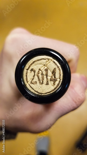 Close up POV shot of a hand holding the neck of a glass wine bottle, displaying the cork with written on it the year of the grape harvest, 2014. 