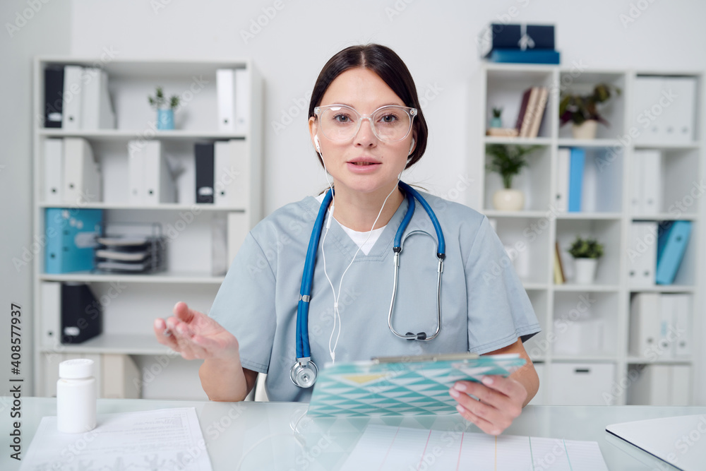Serious young female doctor in uniform and earphones talking to online patient