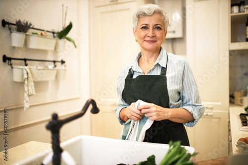 Attractive middle aged woman with short hair doing housework standing at sink wearing apron, holding towel, cleaning kitchen, cooking vegetable meal. Joyful mature female cooking breakfast for family