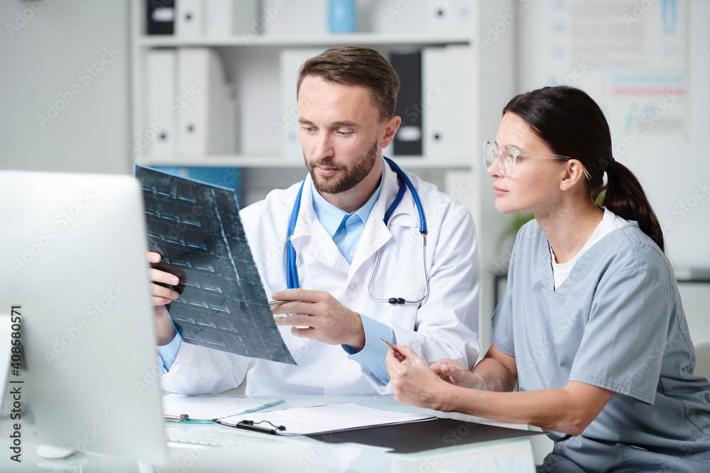 Two young clinicians in uniform sitting by desk and discussing x-ray of patient Two young clinicians in uniform sitting by desk and discussing x-ray of patient