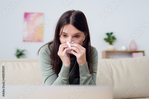 Young sick woman in homewear blowing her nose while sitting in front of laptop