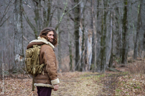 Rear view of a young man with long hair, sheepskin winter coat and green military backpack looking at camera while walking in the woodlands.