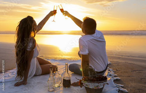 A gentle romantic date on the ocean during the magical sunset. A girl and a guy in white clothes sit on a white bedspread on the ocean during a magical sunset and drink a drink from beautiful glasses.