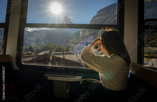 Young Asian woman sitting on train at window seat admires the fantastic landscape of Matterhorn mountain peak with sun in the sky out of window with hand shielding sun and take pictures by cell phone.