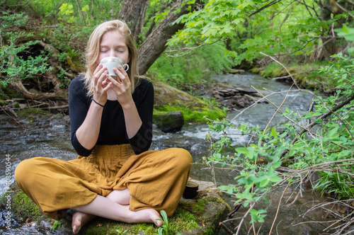 Portrait of a pretty young girl sitting cross legged on a rock on the shore of a river, holding a china tea cup with both hands and drinking