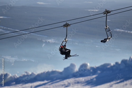 Persons sitting in a ski lift with snow in the fore ground and a wide landscape in the background