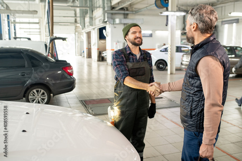 Happy young bearded technician of car service shaking hand of male client