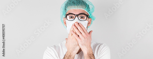 Doctor surgeon in white medical mask and a medical cap stands over white background with surprise,covers his mouth with the hands. Сoncept of covid 19, flu and seasonal cold.