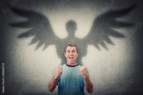 Courageous middle aged man keeps fists tight raised up, screaming showing his powers. Confident male casting a superhero shadow with angel wings on the wall. Inner strength and ambition concept photo