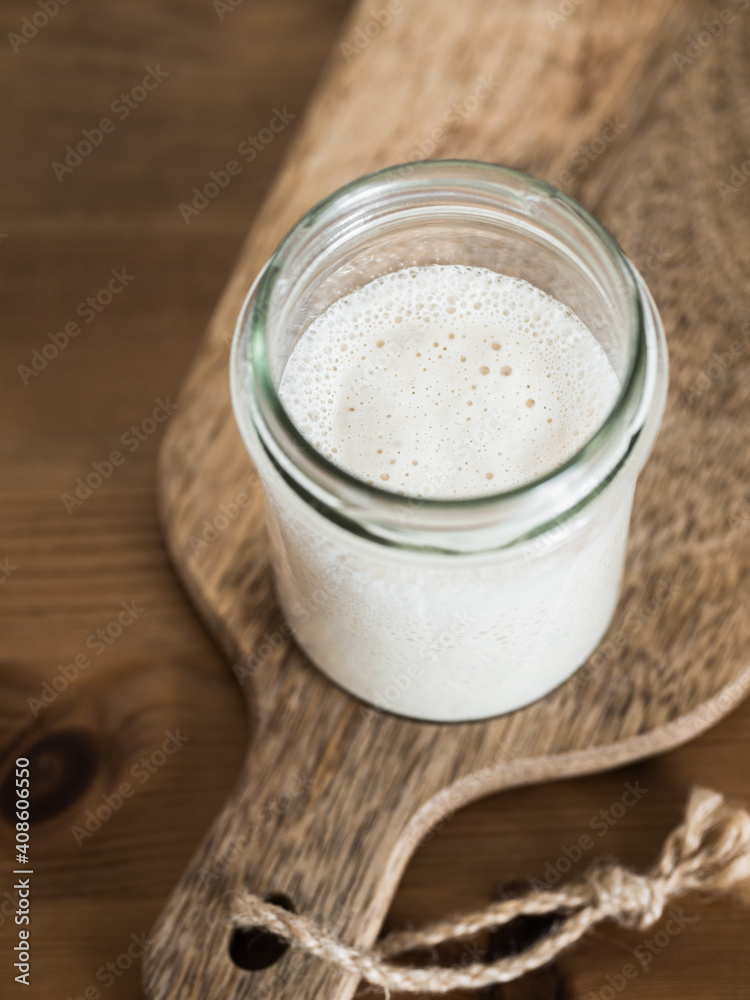 Wheat sourdough starter. Glass jar with sourdough starter on wooden cutting board over wooden tabletop. Copy space for text or design. Vertical. Wheat sourdough starter. Glass jar with sourdough starter on wooden cutting board over wooden tabletop. Copy space for text or design. Vertical.