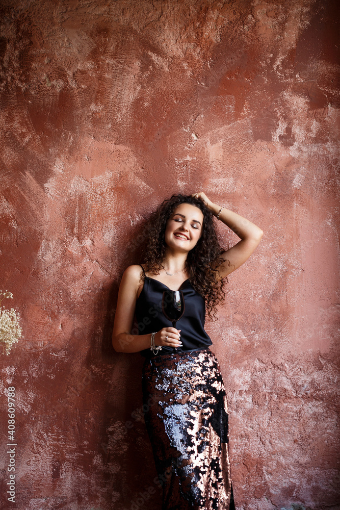 Portrait of a young curly woman of European appearance against the background of a red wall, dressed in a black satin top and skirt