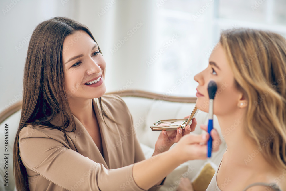 Dark-haired smiling girl doing make up to her friend Dark-haired smiling girl doing make up to her friend