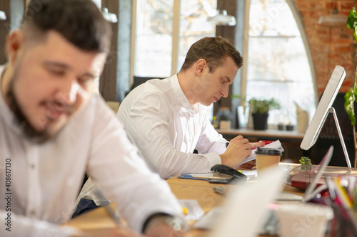 Team project, inspired. Young caucasian colleagues working together in a office using modern devices and gadgets. Look busy, attented, cheerful, successful. Concept of business, office, finance.