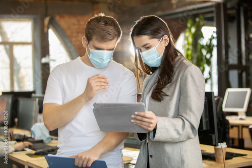 Young caucasian colleagues working together in a office using modern devices during quarantine. Look busy, attented, cheerful, successful. Concept of business, office, finance. Wearing face masks