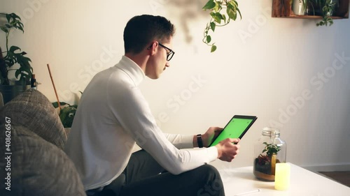 Young Man Sitting on a sofa Using Tablet Smart Device with Green Mock-up Screen in vertical Mode. Business Man Using Application, Website, Content. Social Media, E learning Course in the evening. photo