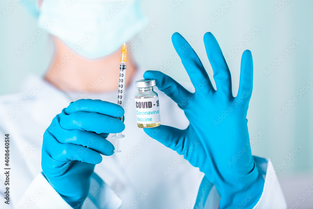 Woman doctor or nurse in uniform and gloves wearing face mask protective in lab holding medicine vial vaccine bottle with COVID-19 Coronovirus vaccine label Woman doctor or nurse in uniform and gloves wearing face mask protective in lab holding medicine vial vaccine bottle with COVID-19 Coronovirus vaccine label