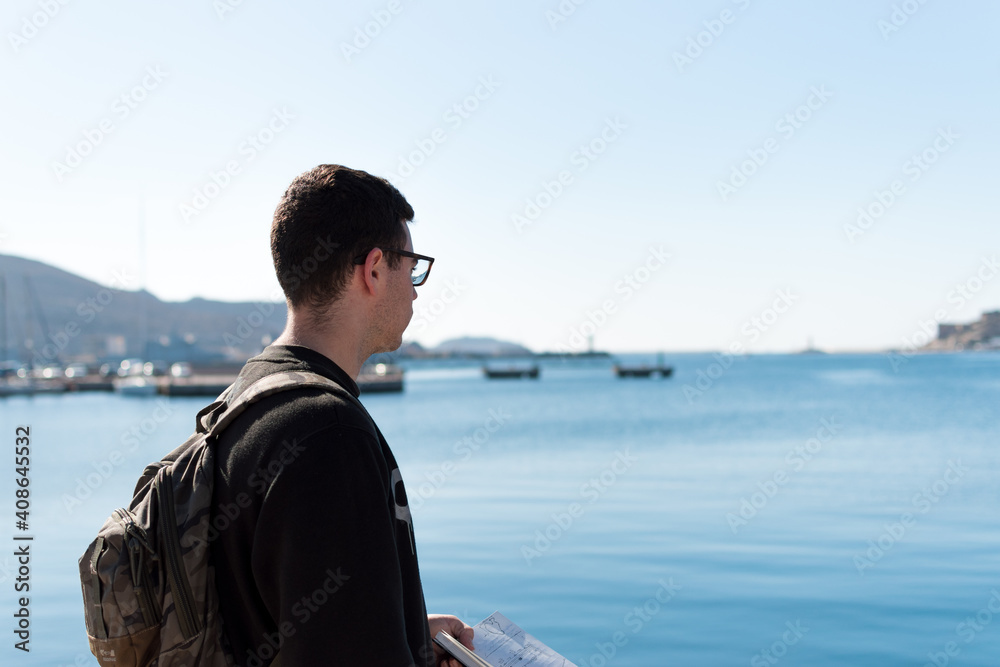 young male 20s student on his back wearing a black sweatshirt, a militar backpack and glasses holding a notebook and looking to the sea young male 20s student on his back wearing a black sweatshirt, a militar backpack and glasses holding a notebook and looking to the sea