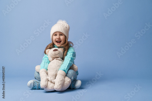 happy little 5-year-old girl with blonde hair in a knitted sweater cap and mittens, smiles and hugs teddy bear sitting on blue background in the studio. toy is her friend. Winter time. Holiday weekend