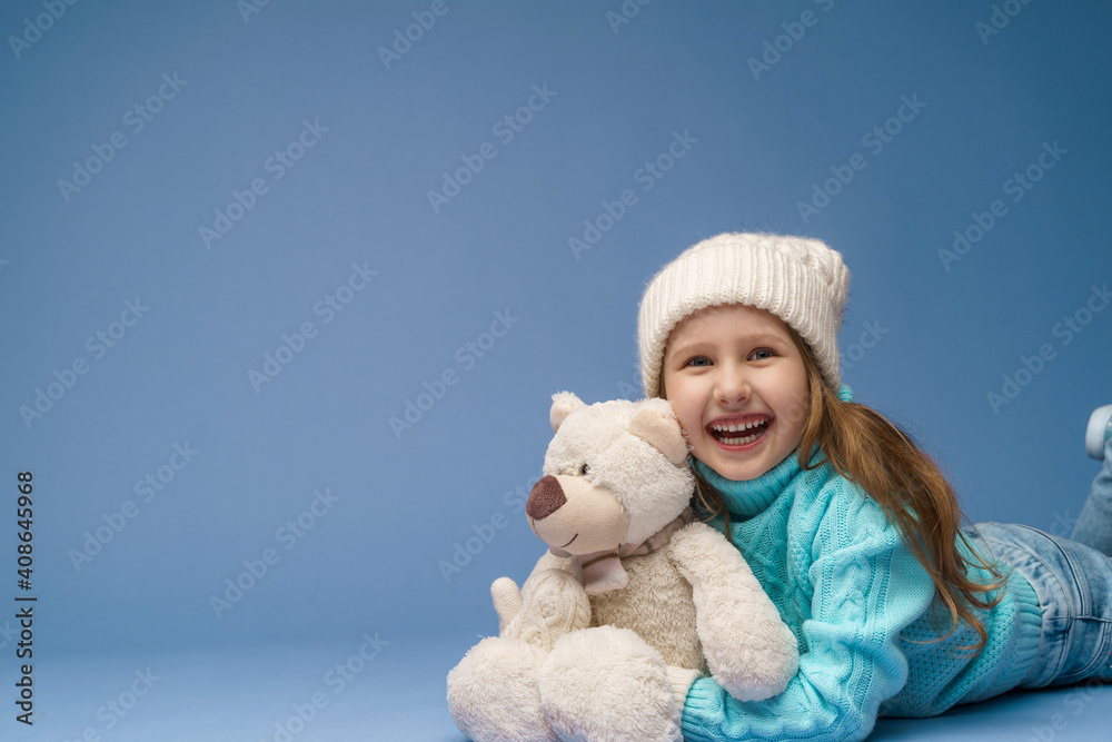 happy little 5-year-old girl with blonde hair in a knitted sweater cap and mittens, smiles and hugs teddy bear lying on blue background in the studio. toy is her friend. Winter time. Holiday weekend