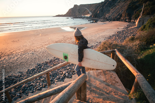 Young woman in casual clothes walk down stairs to beautiful epic beach at sunset or sunrise. Female surfer ready to start surfing lesson at dawn. Millennial lifestyle and hobby. Cinematic wanderlust photo