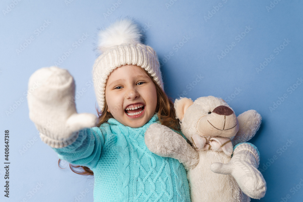 happy little 5-year-old girl with blonde hair in a knitted sweater cap and mittens, smiles and hugs teddy bear lying on blue background in the studio. toy is her friend. Winter time. Holiday weekend happy little 5-year-old girl with blonde hair in a knitted sweater cap and mittens, smiles and hugs teddy bear lying on blue background in the studio. toy is her friend. Winter time. Holiday weekend