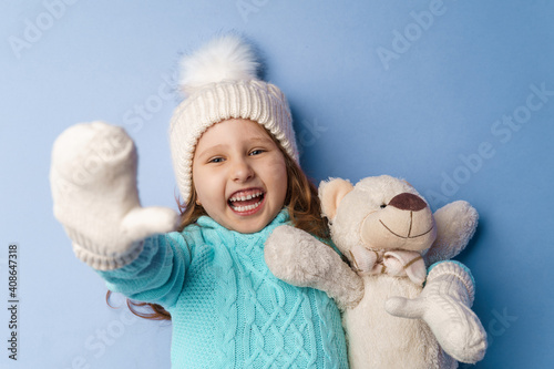 happy little 5-year-old girl with blonde hair in a knitted sweater cap and mittens, smiles and hugs teddy bear lying on blue background in the studio. toy is her friend. Winter time. Holiday weekend