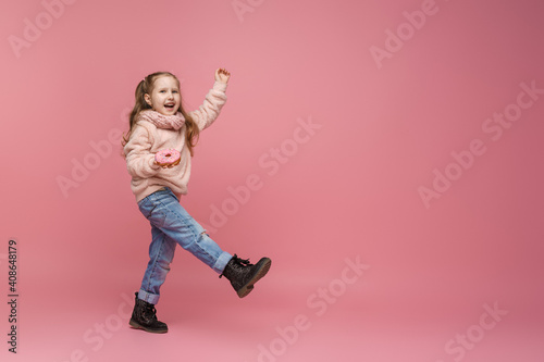 little girl in a fluffy sweater and a headband, smiling with a doughnut in her hand. Sweet appetizer, delicious pastries. a child shows a delicious dessert on a pink background in studio