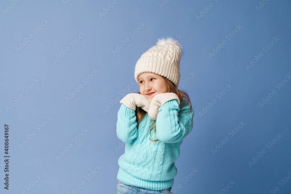 cute little girl in white knitted hat, sweater and mittens has her hands pressed to her face and is smiling against blue background. child in warm clothes looks away. Advertising. Winter. Copy space cute little girl in white knitted hat, sweater and mittens has her hands pressed to her face and is smiling against blue background. child in warm clothes looks away. Advertising. Winter. Copy space