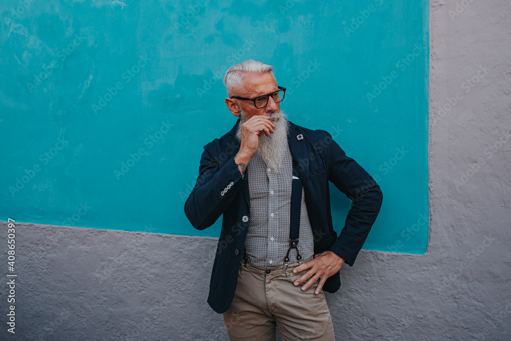 an older man in hipster clothes and glasses and a long white beard poses on a blue wall an older man in hipster clothes and glasses and a long white beard poses on a blue wall