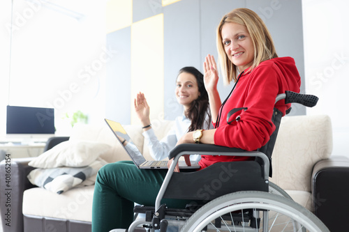 Woman in wheelchair with laptop on her lap waving in greeting with her girlfriend. Remote work for people with disabilities health concept