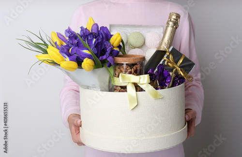 Woman holding basket with gifts, bouquet and champagne on light grey background, closeup photo