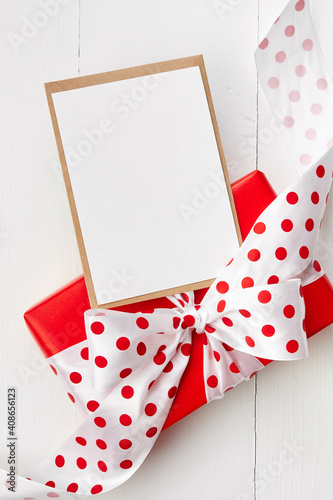 Greeting card mockup and red gift box with big bow on white wooden background